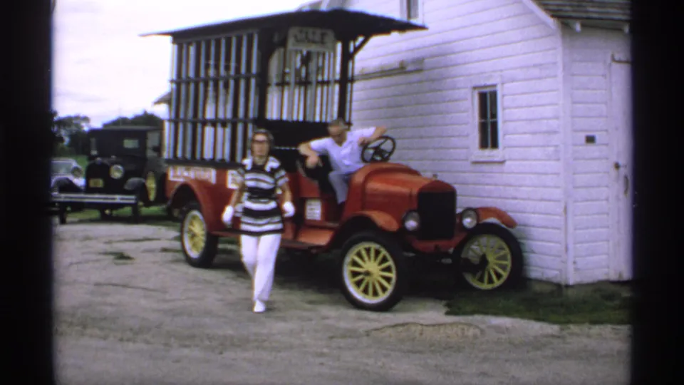 The woman walks in front of the red antique-style sales car, with the man still seated beside the white clapboard building.