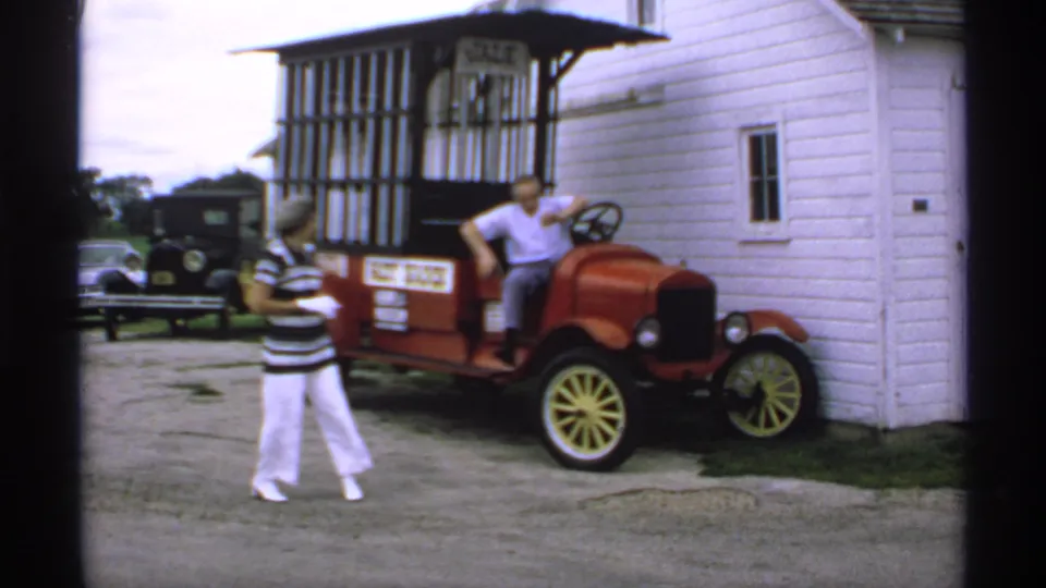 At the end of the 1970 Minnesota clip, the woman crosses the foreground and the man remains seated in the red sales car by the white building.