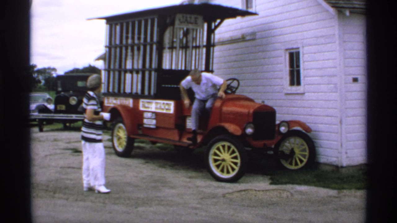 Woman watches as a man climbs into a red antique-style car beside a white building.