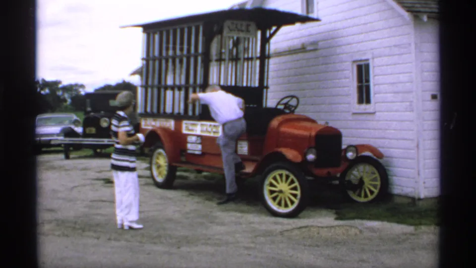 1970 Minnesota home-movie frame with a man leaning into a red antique-style vehicle as a woman observes near a white building.