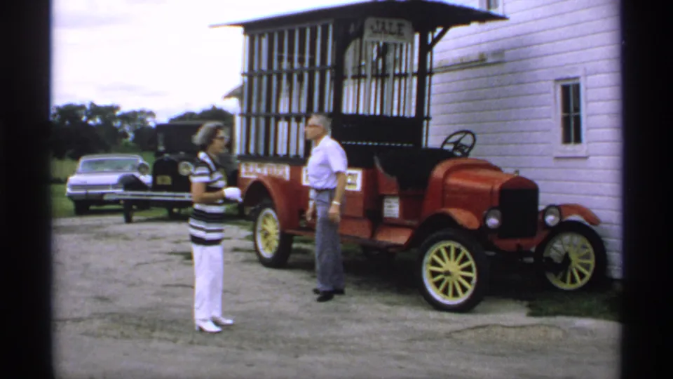 1970 Minnesota frame showing two adults facing each other near a red antique-style car, with parked cars visible behind them.