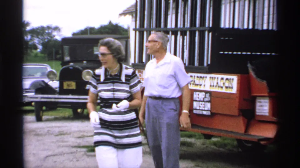1970 Minnesota 8mm frame of a woman and man walking beside a red antique-style display car, with older parked cars in the background.