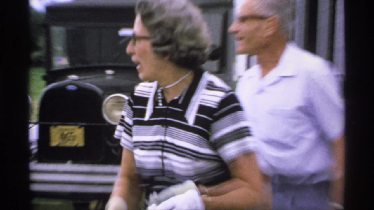 Woman in striped dress and white gloves walks beside a man near a black classic car.