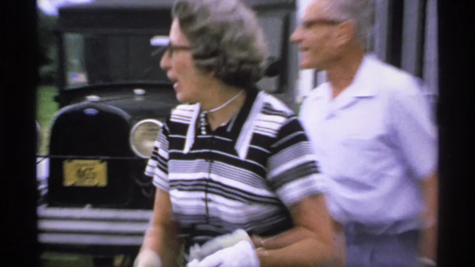 1970 Minnesota home-movie frame with two adults walking near a black classic car and a white-sided building.