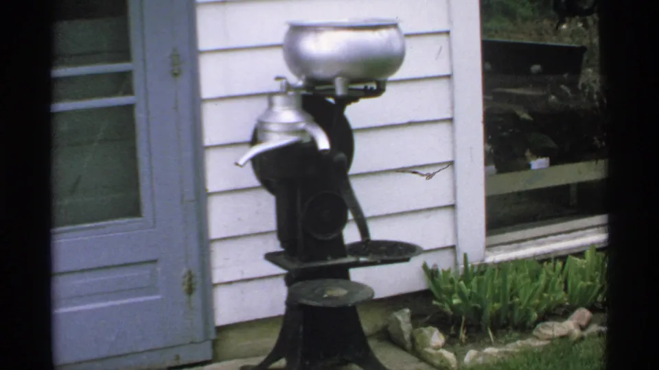 1970 Minnesota home-movie frame showing an antique milk separator outside a white house with a blue door.