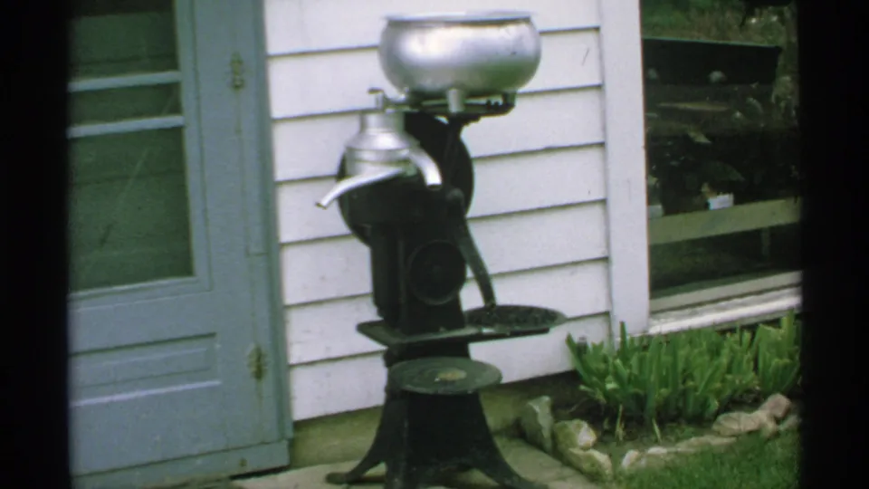 1970 Minnesota home-movie frame with a closer handheld view of an outdoor antique milk separator by a house.