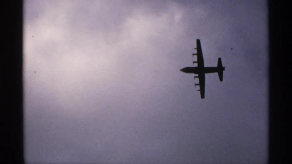 A dark four-engine propeller plane crosses a pale cloudy sky in 1970 Minnesota home-movie footage.