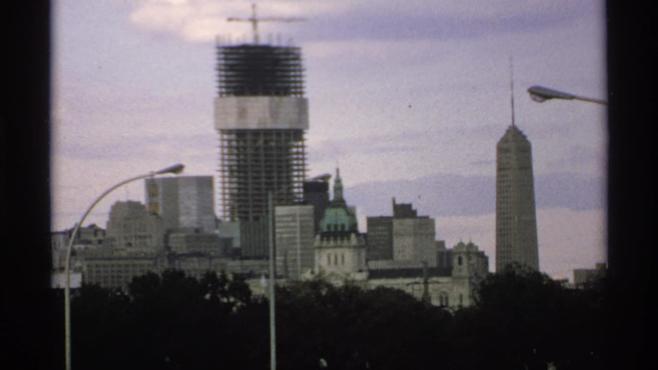Distant Minnesota skyline with a high-rise under construction, spired tower, treetops, and streetlights.