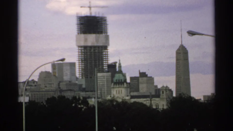 1970 8mm view of a Minnesota downtown skyline with a crane-topped tower under construction, older buildings, treetops, and streetlights.