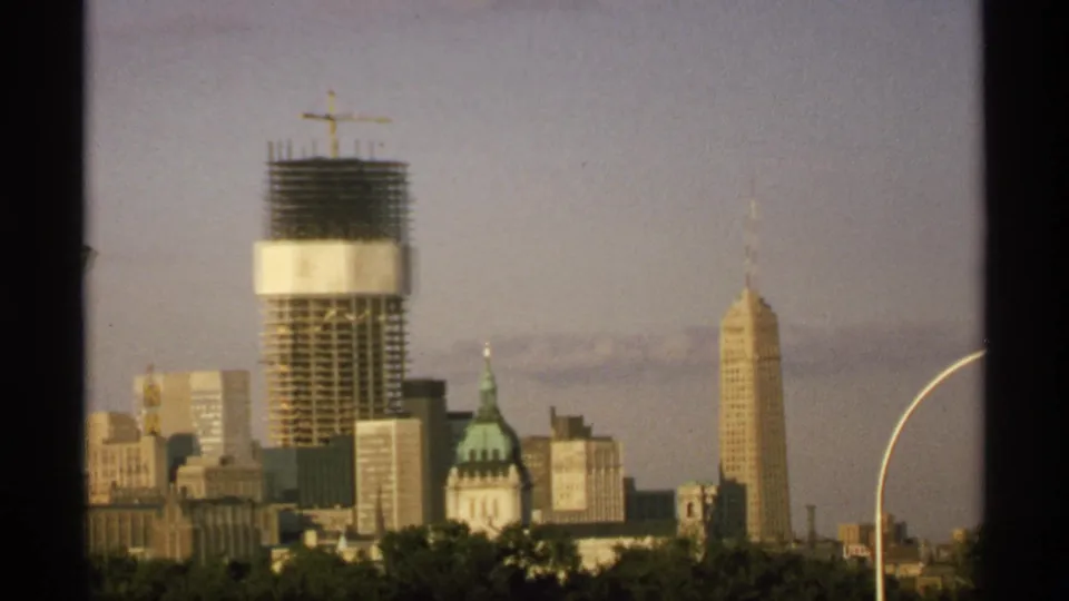 1970 Minnesota skyline view showing a sunlit tower under construction, a green-domed building, a spired high-rise, trees, and open sky.