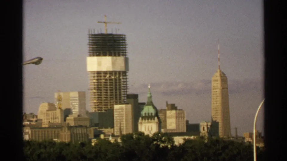 1970 8mm frame of a Minnesota downtown skyline, centered on a tower under construction with a crane and nearby spired and domed buildings.