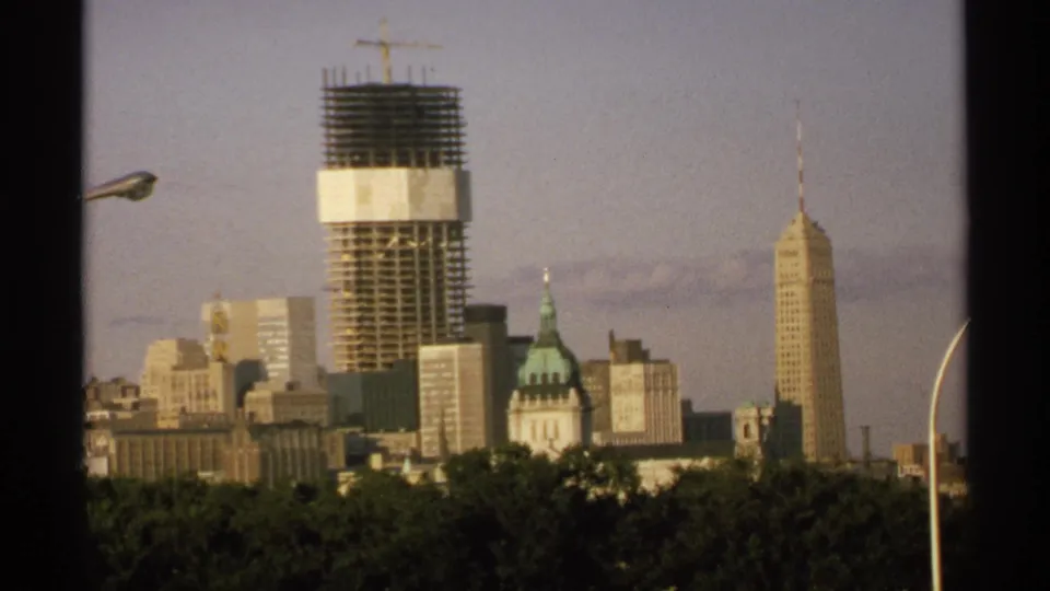 Wide 1970 Minnesota city view on 8mm film with a crane-topped high-rise, spired tower, domed building, treetops, and curved streetlights.