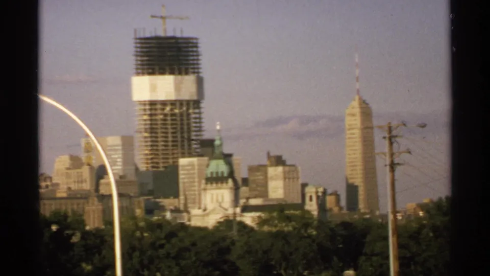 1970 Minnesota skyline frame with modern buildings, a tower under construction, green-domed structure, spired high-rise, trees, streetlight, and utility pole.