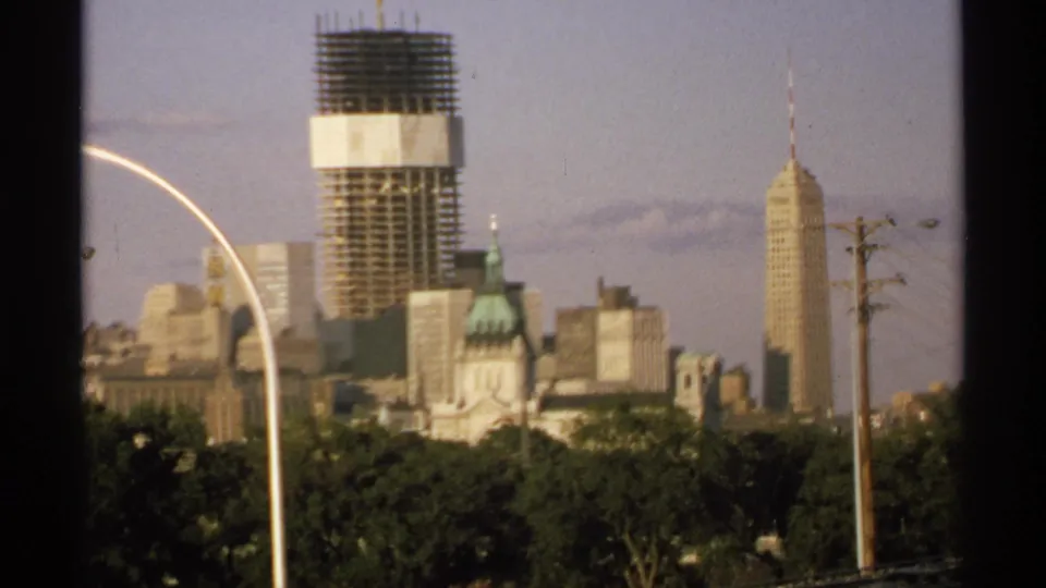 1970 8mm Minnesota city view with a softened skyline, high-rise construction, green dome, spired tower, roadside streetlight, utility poles, and trees.