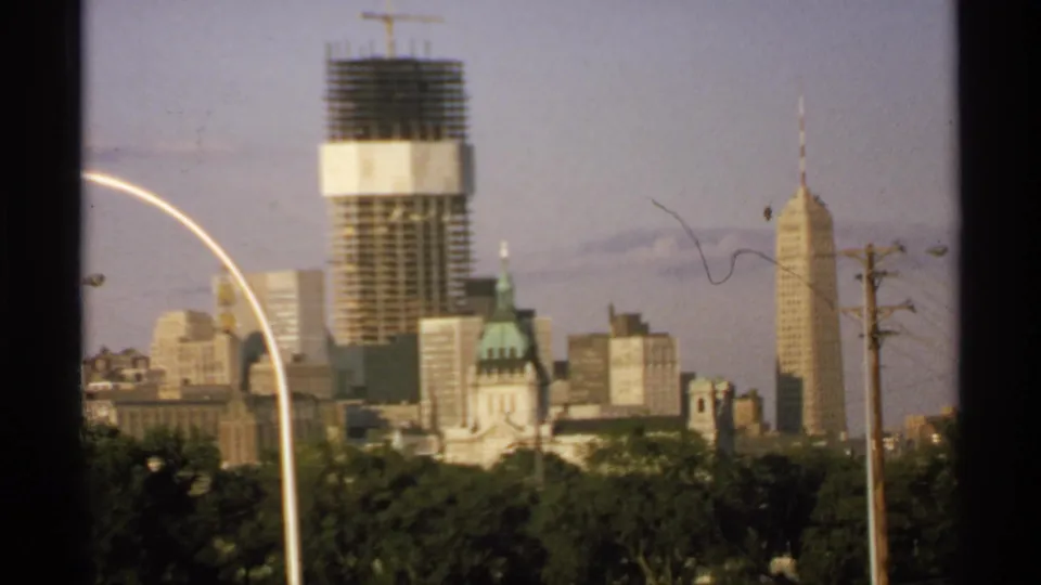 Final 1970 Minnesota skyline frame showing a construction high-rise, green-domed building, spired tower, trees, utility poles, and warm 8mm film color.