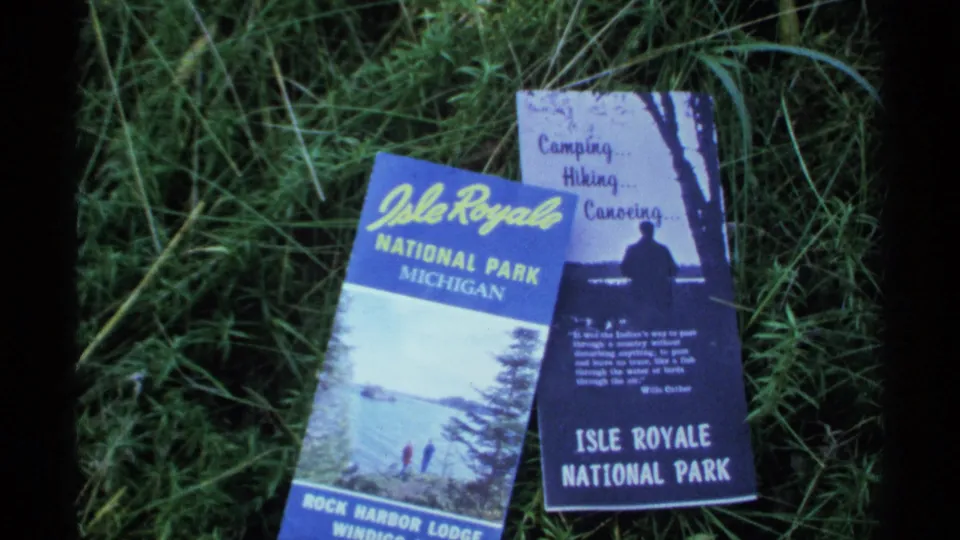 Two vintage Isle Royale National Park brochures rest in grass, one showing Rock Harbor Lodge text and another listing camping, hiking, and canoeing.