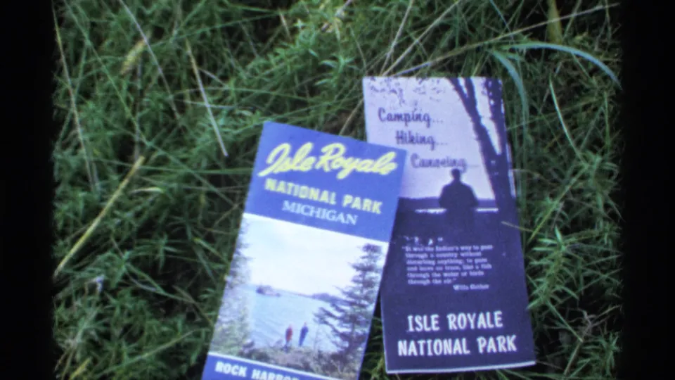 1971 8mm frame showing Isle Royale National Park brochures on sunlit grass during a Michigan vacation.