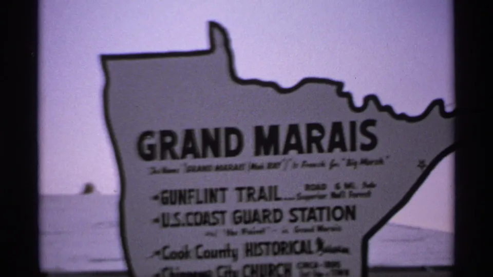 The Grand Marais sign fills the frame, with water and distant shoreline visible behind the roadside marker.