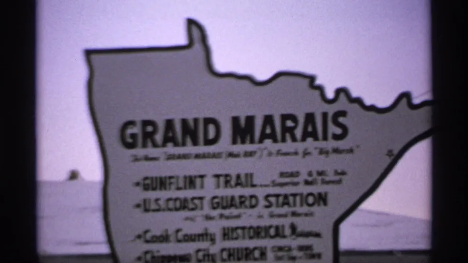 1971 8mm frame of a Grand Marais sign listing local points of interest, filmed with soft focus near the waterfront.