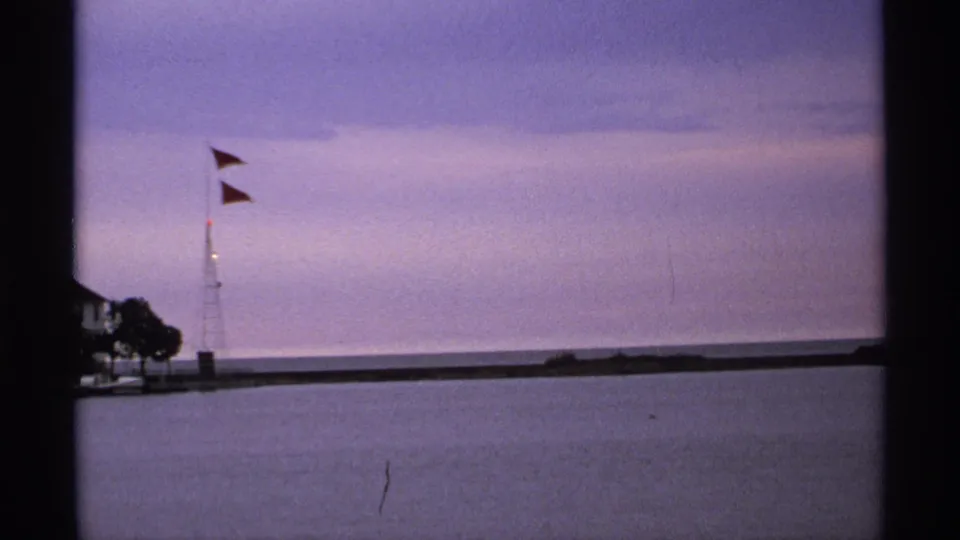 Quiet 1971 lakeshore view with red flags flying near a small harbor light or mast and a long breakwater under a muted sky.