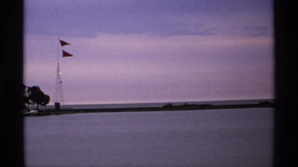 Home-movie harbor shot with two red flags on a mast, a low breakwater, and still lake water in a misty Great Lakes setting.