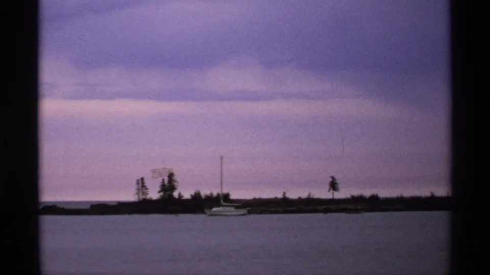 1971 8mm waterfront frame showing a small sailboat near a dark tree-lined shore beneath a purple-tinged sky.