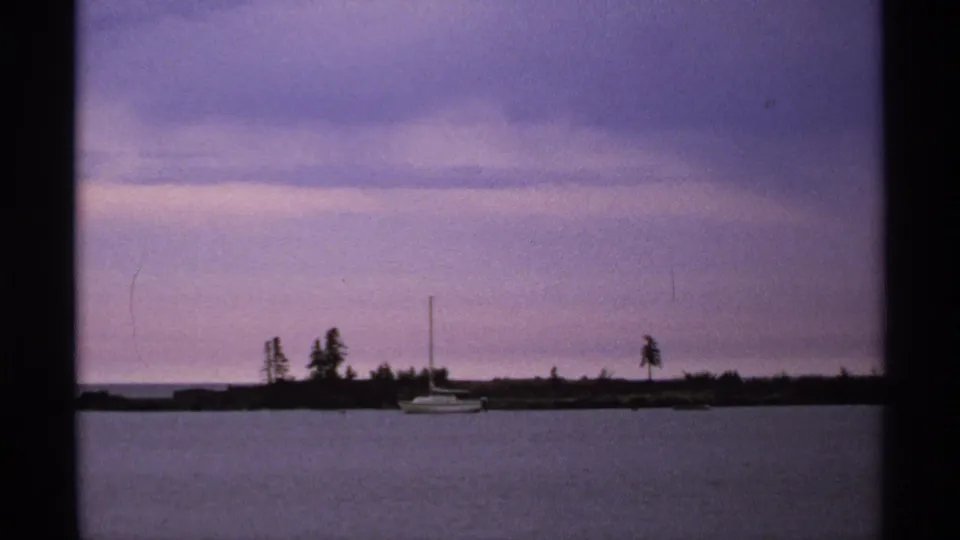 1971 8mm Isle Royale view of a small sailboat floating near a wooded lakeshore beneath a muted dusk sky.