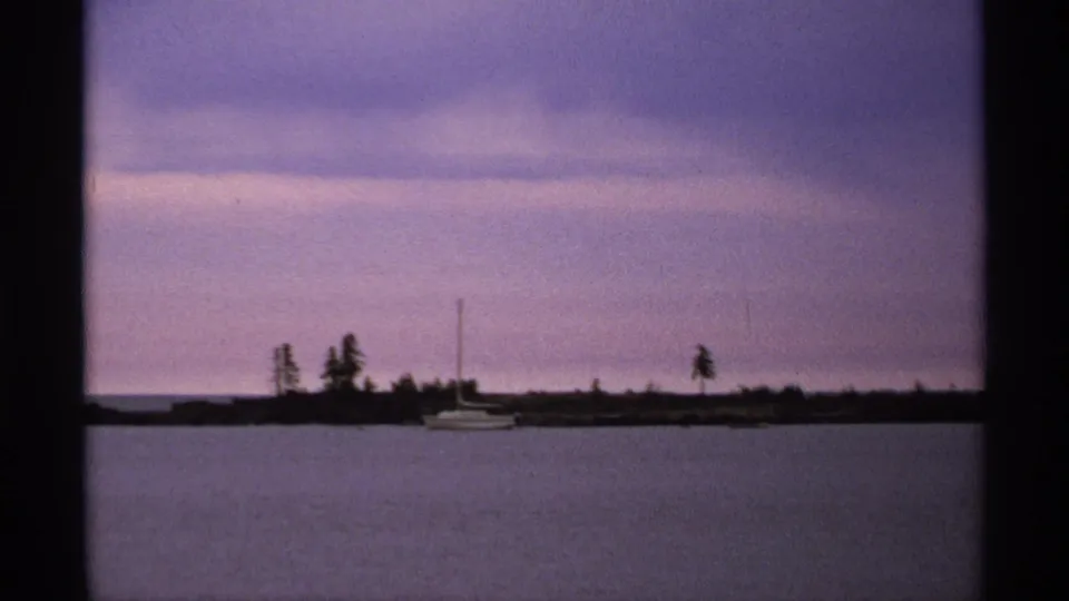 1971 Isle Royale home-movie frame showing a sailboat near a tree-lined shore across calm water at dusk.