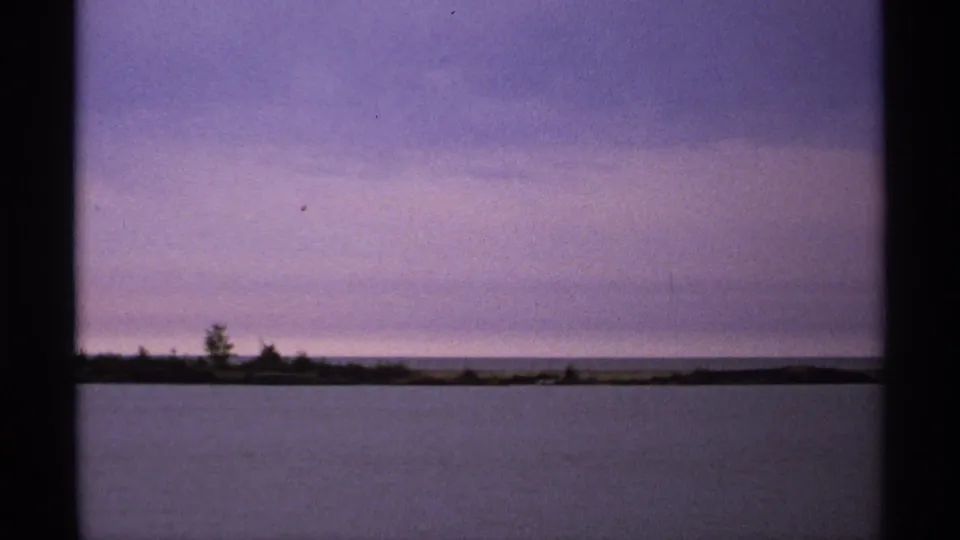 1971 Isle Royale lakeshore frame with still water, sparse trees, and a subdued purple dusk sky.