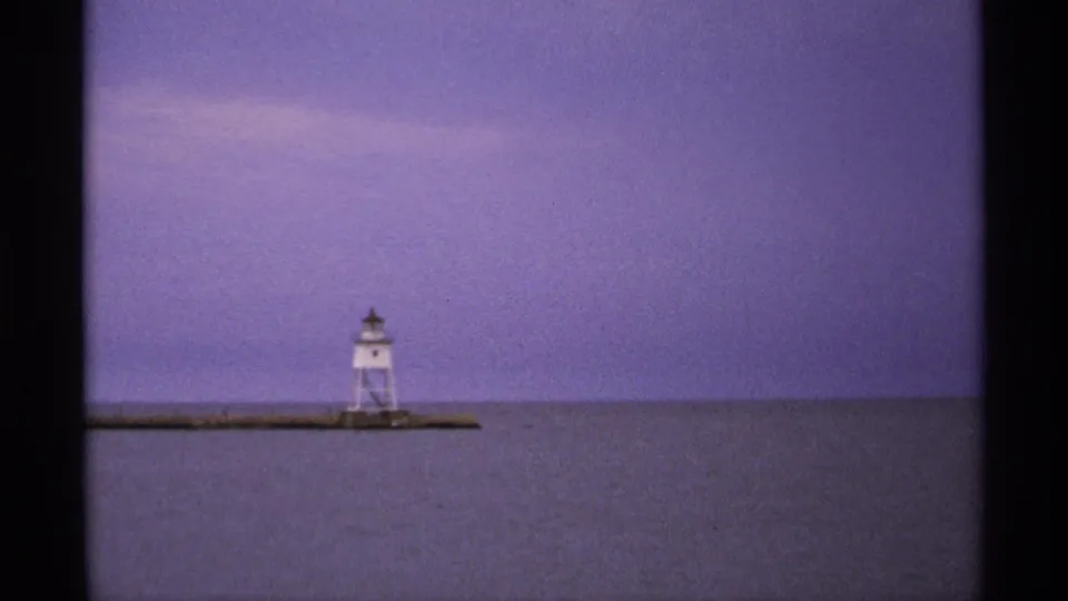 1971 Isle Royale 8mm view of a small white lighthouse on a pier against calm lake water and a violet dusk sky.
