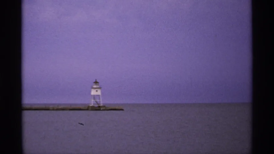Isle Royale 8mm frame showing a white lighthouse on a narrow pier with still water and a purple evening sky.