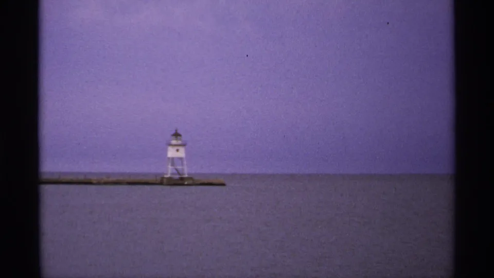1971 Isle Royale home-movie frame showing a distant white tower on a low breakwater across Lake Superior water.