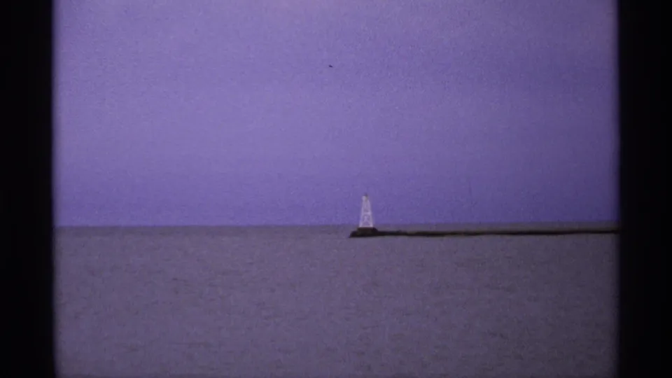 1971 8mm Isle Royale frame with a distant narrow tower on a breakwater across calm Lake Superior water.