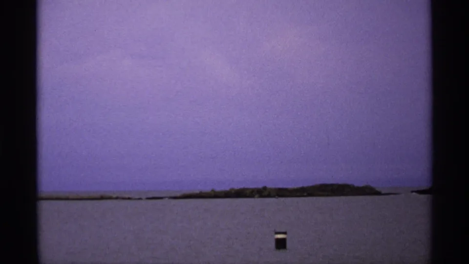 1971 Isle Royale home-movie frame showing a distant island shoreline across water and a small dark object in the foreground.