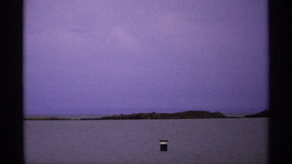 Isle Royale shoreline viewed from across the water in 1971, with a dark floating object and hazy sky visible.