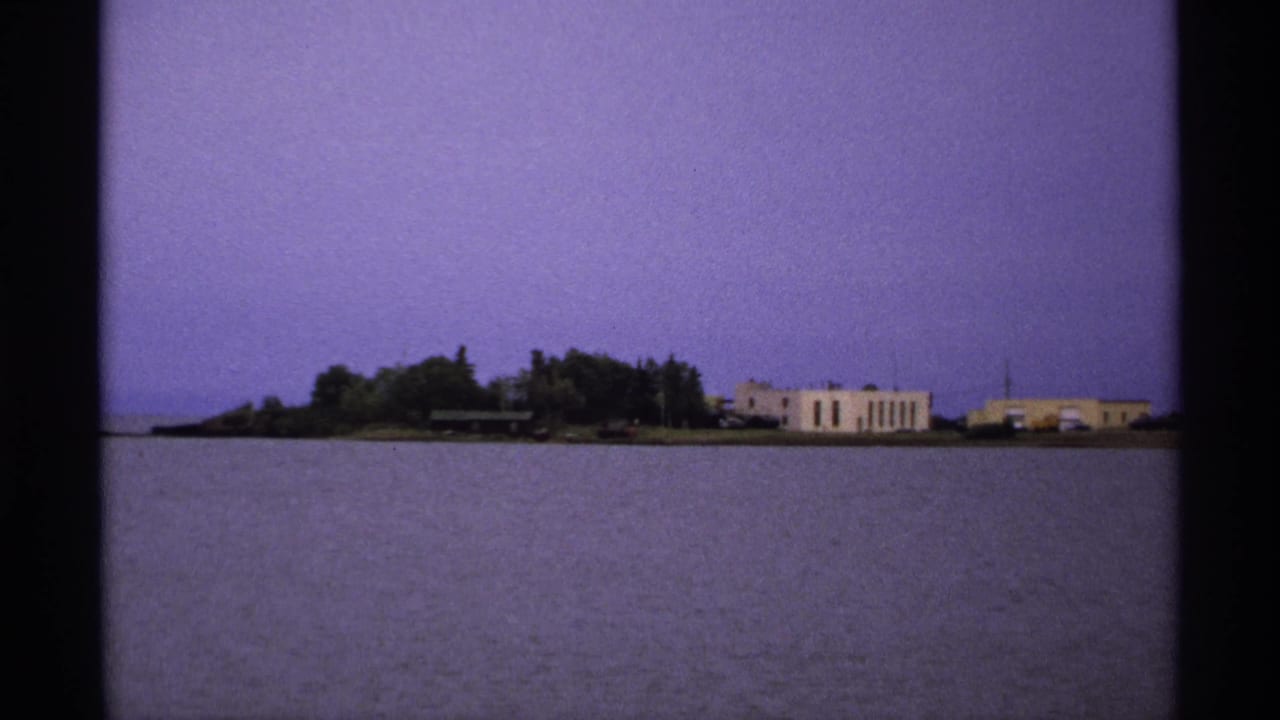 1971 Isle Royale 8mm frame showing calm lake water, a distant wooded shoreline, and low lakeside buildings under a purple-tinted sky.