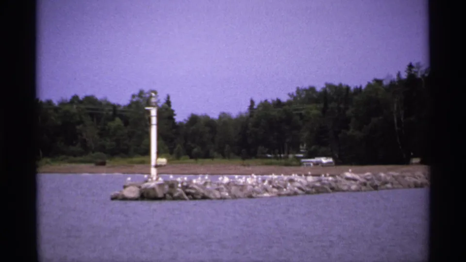 1971 Isle Royale 8mm frame of a rocky lakeside jetty with a white pole or beacon, wooded shoreline, and birds perched on the rocks.