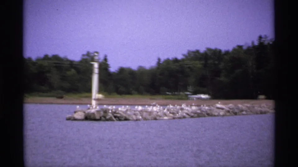 1971 Isle Royale 8mm frame centered on a rocky breakwater with a white pole, perched birds, lake water, and a wooded shoreline.
