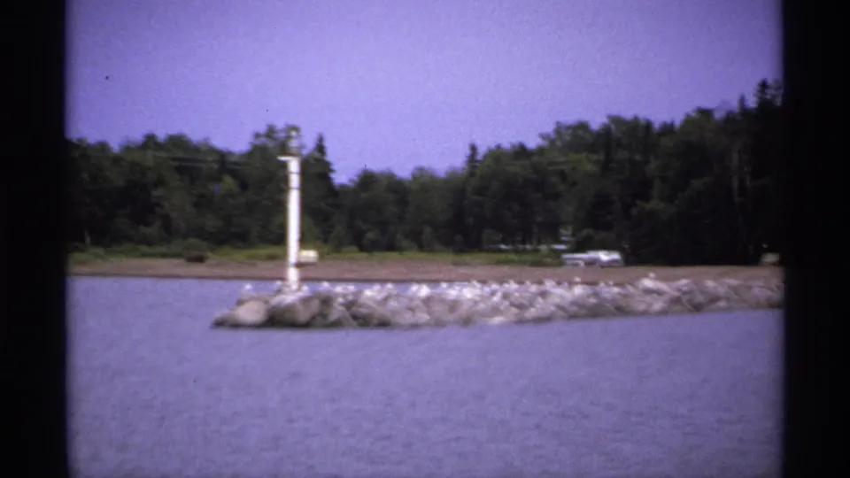 1971 Isle Royale 8mm frame showing the jetty from a slightly shifted angle, with a white pole, birds, water, and forested shore.