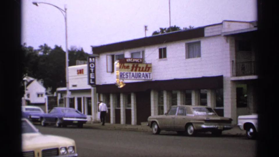The Hub Restaurant and Motel remain in view as a man walks along the sidewalk past parked cars in this 1971 home-movie frame.