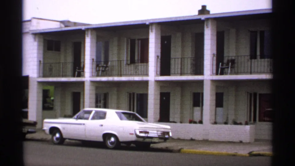 A two-story motel building with balcony railings and room doors is shown, with a white period car parked along the curb.
