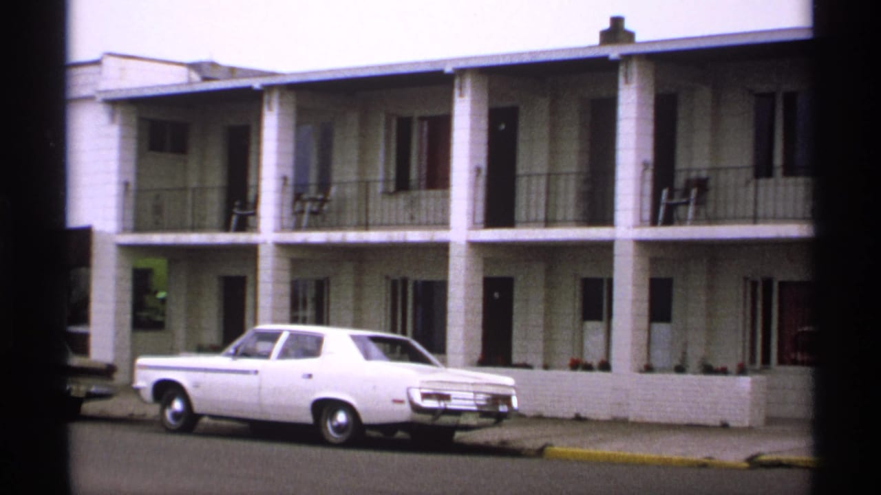 1971 Isle Royale home-movie frame showing a white sedan outside a two-story lodging building with balcony railings and dark film edges.