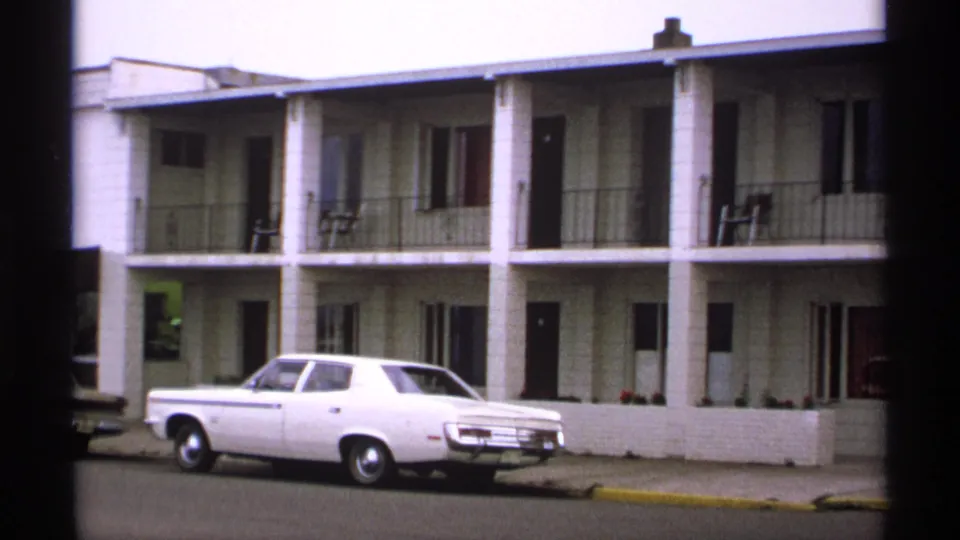 1971 Isle Royale home-movie frame showing a white sedan outside a two-story lodging building with balcony railings and dark film edges.