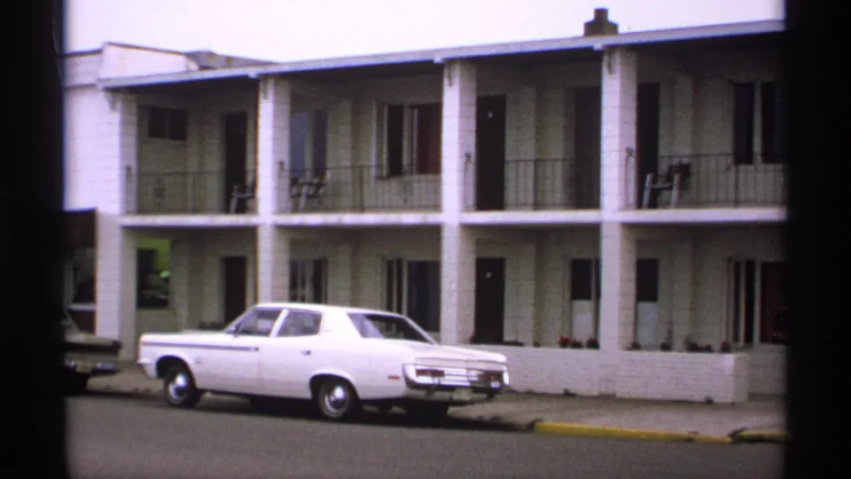 1971 Isle Royale frame of a white classic sedan parked by a motel-style lodging exterior with railings, doors, and curb visible.