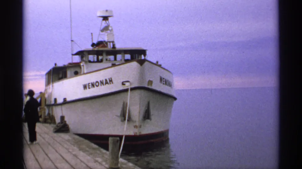 1971 Isle Royale waterfront frame showing the white boat WENONAH tied at a wooden pier, with calm water and a silhouetted person nearby.