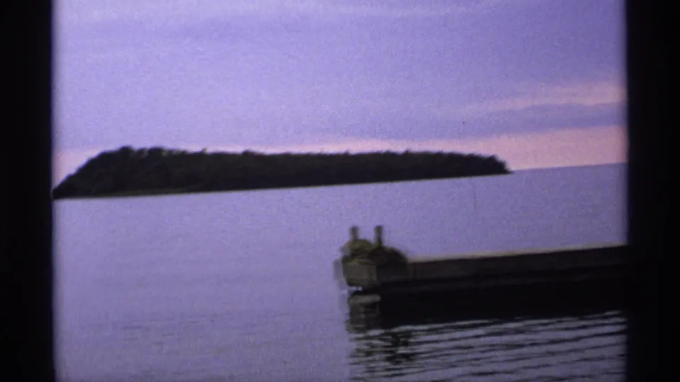 Isle Royale home-movie frame with a dark island silhouette across calm lake water and a dock edge in the foreground.