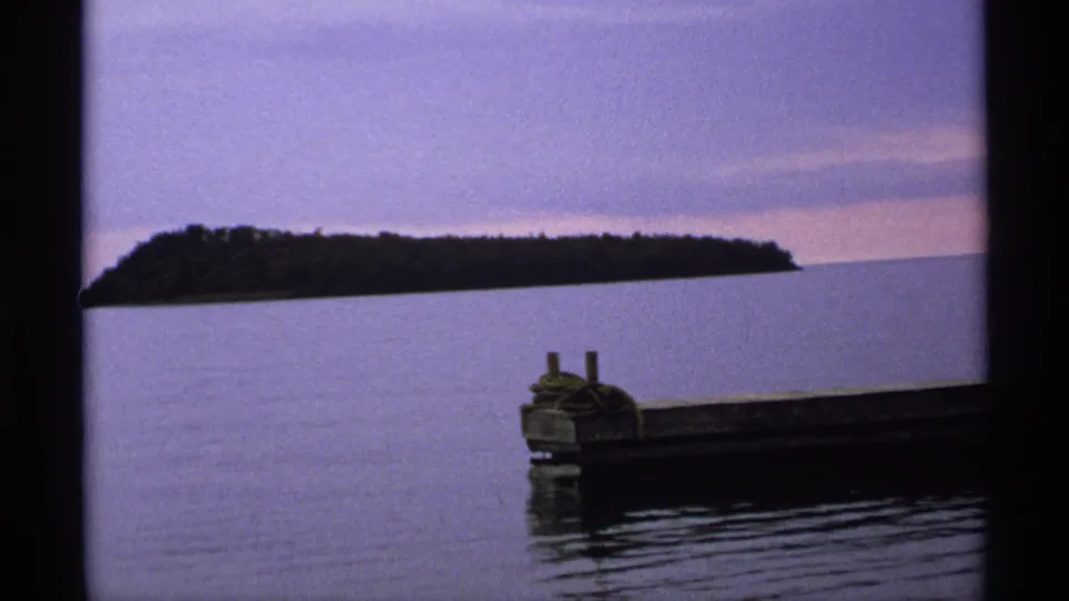1971 Isle Royale frame showing a long wooded island silhouette beyond still lake water, with a dock cutting across the foreground.
