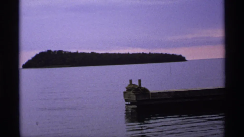 Isle Royale 8mm frame of a low wooded island across the lake, framed by a dock structure at lower right under a dim sky.