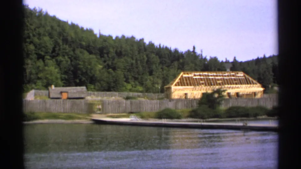 1971 Isle Royale frame showing a wooden palisade, exposed roof framing, dock, small boat, and dense forest along the lakeshore.