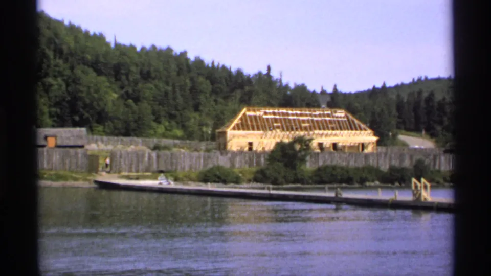 1971 Isle Royale home-movie frame with a dock, palisade wall, exposed wooden roof frame, small boat, and forested slope behind the site.